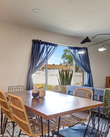 Open-plan dining and living room with wood table, rattan chairs, blue curtains, and desert view through glass doors