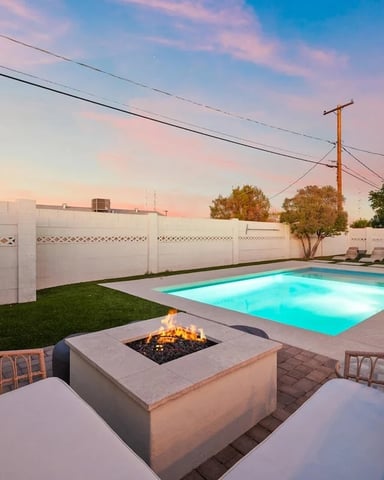 Backyard with illuminated swimming pool, fire pit, lounge chairs, and white privacy fence at dusk