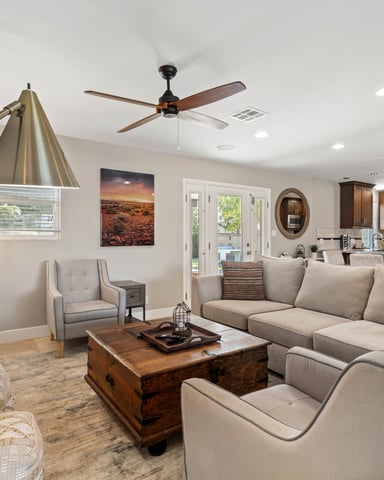 Open-plan living room with beige sectional sofa, wooden coffee table, ceiling fan, and kitchen visible in background