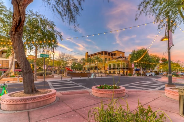 Downtown plaza at dusk with large trees, string lights, terracotta planters, and Spanish colonial-style buildings