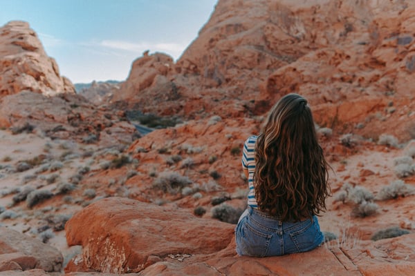 Woman sitting on red rock formation overlooking desert canyon landscape with striped shirt and long dark hair