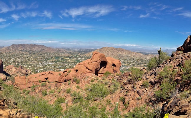 Desert hiking trail through red rock formations with valley and distant mountains under blue sky