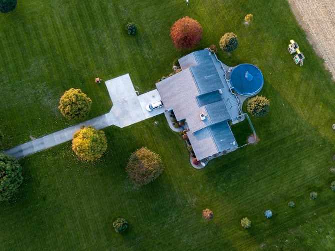 Aerial view of residential home with driveway, pool, and landscaped lawn with scattered trees